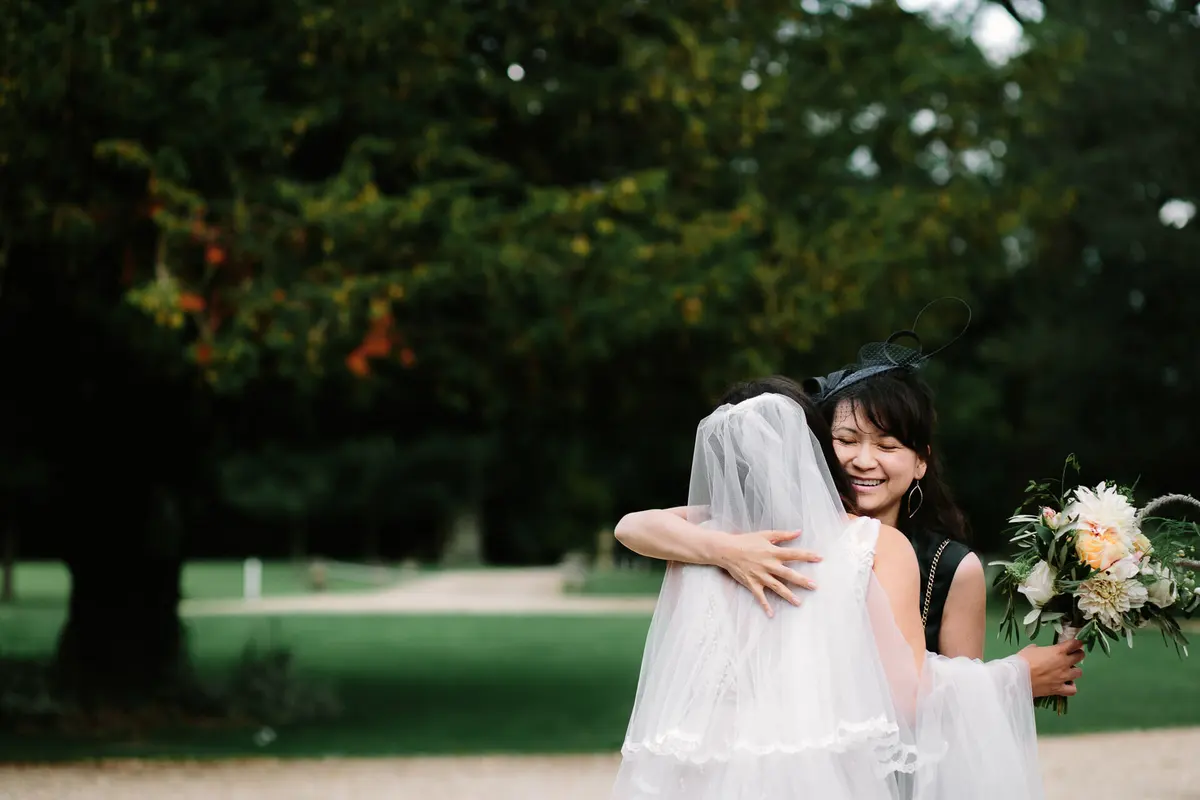 Bride in a long veil hugging a smiling guest in a fascinator on a tree-lined drive