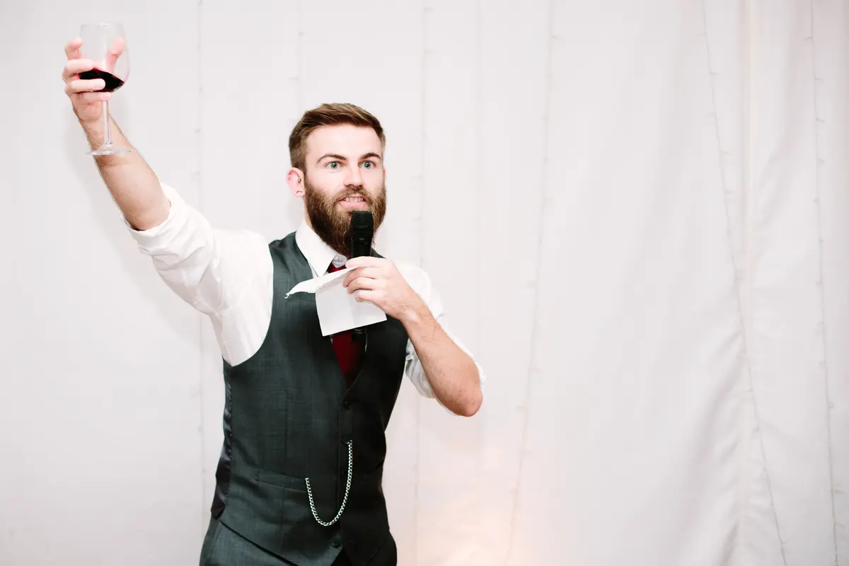 Bearded man in a waistcoat raising a glass of red wine as he gives a speech