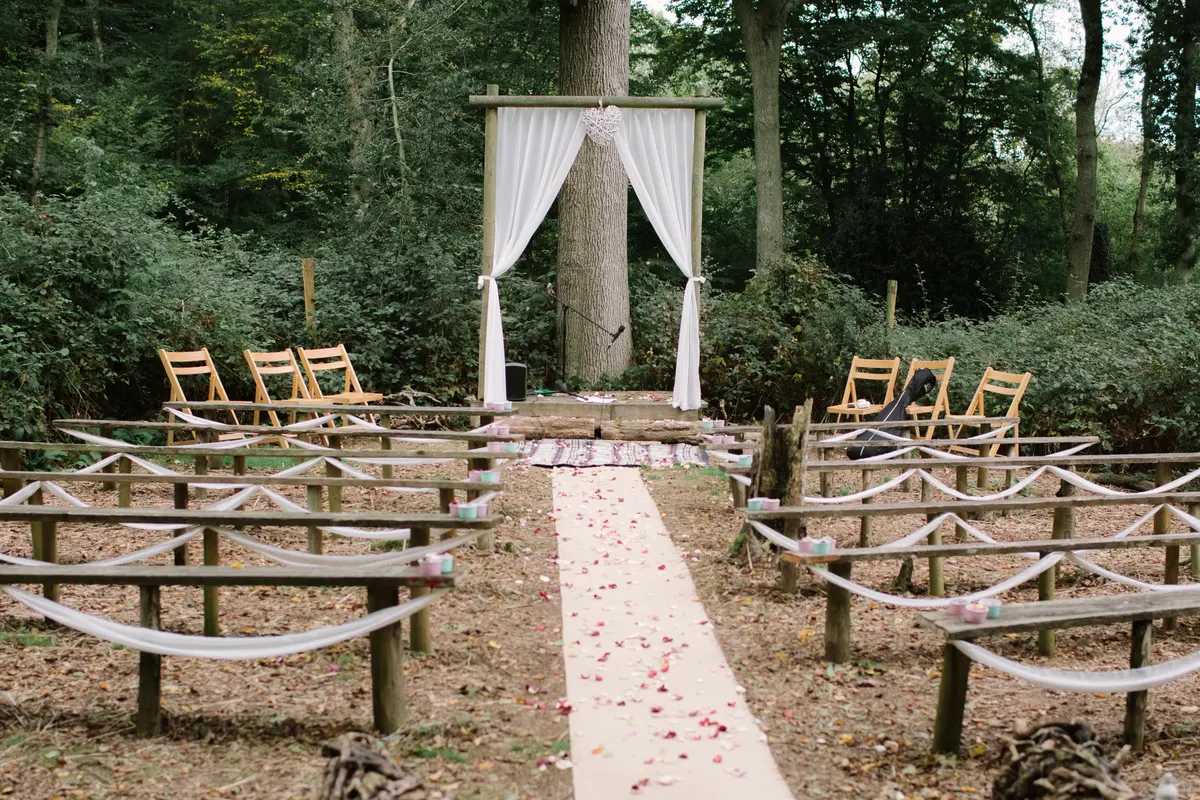Woodland ceremony set-up with rustic benches, a petal-strewn aisle and a draped wooden arch
