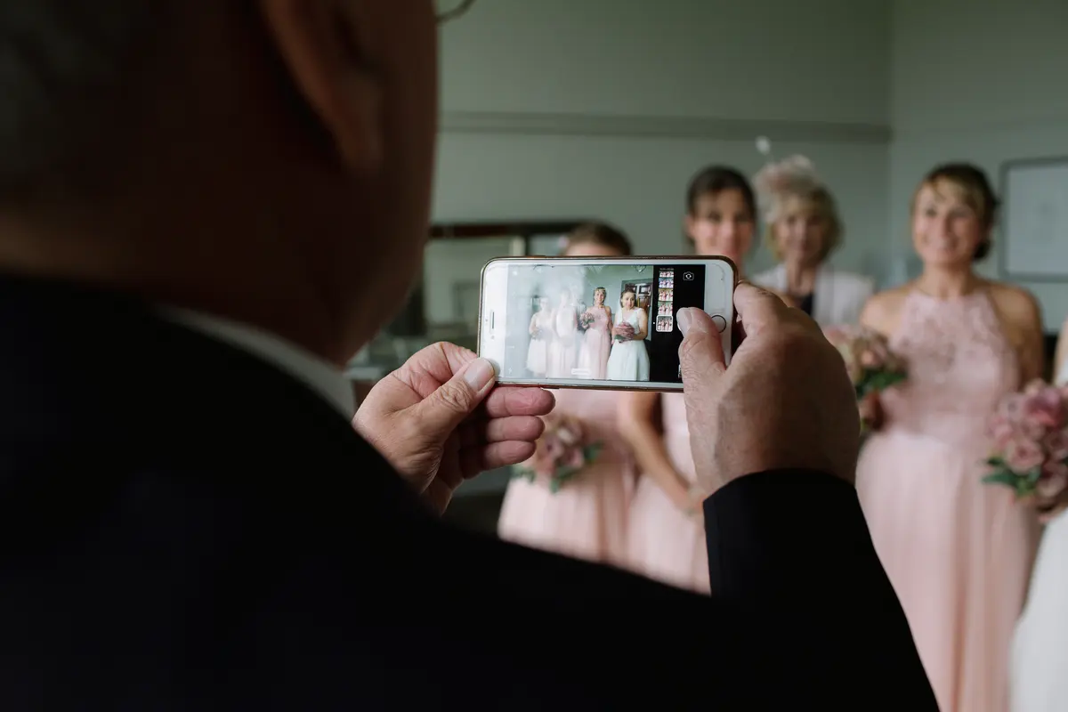 Older man taking a phone photo of bridesmaids in blush pink dresses holding pink roses