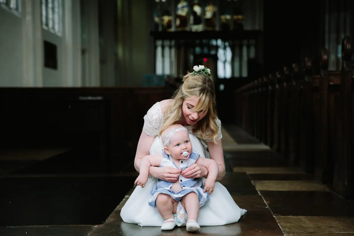 Bride kneeling in a church aisle holding her baby in a blue romper and headband