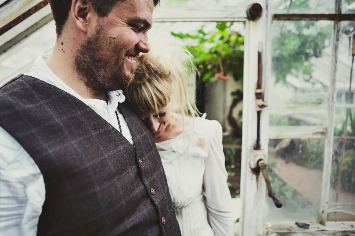 Bride resting her head on the smiling groom's shoulder inside an old greenhouse