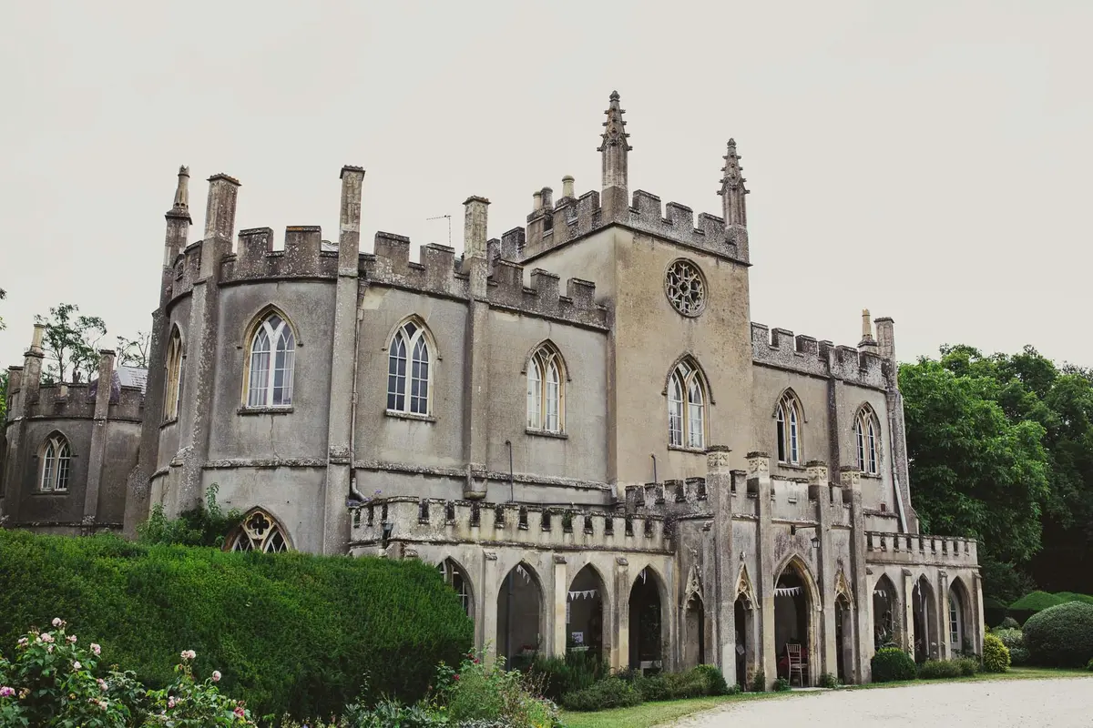 Grey Gothic house with battlements and arched windows behind a tall hedge
