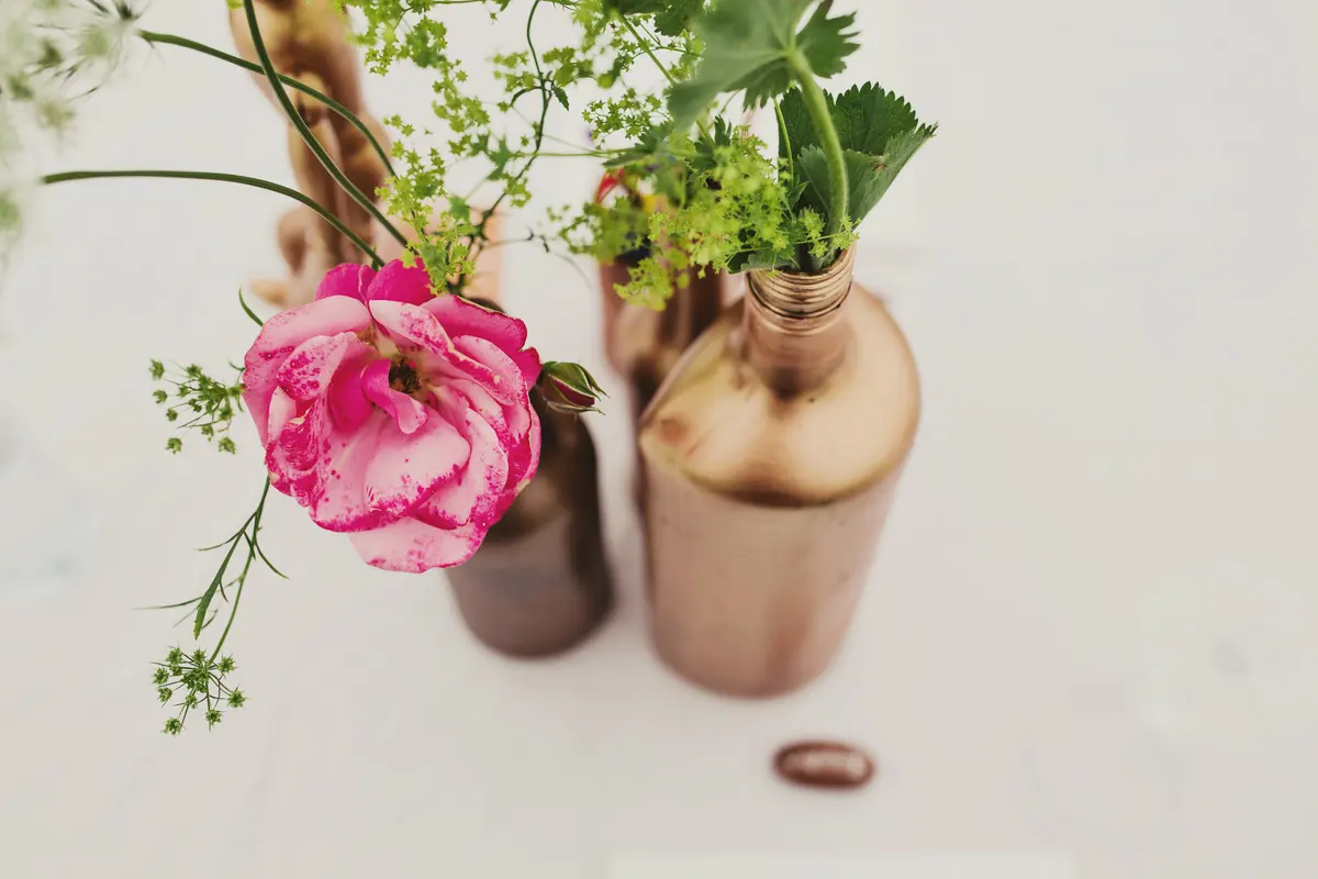 Pink rose and wild flowers in copper-painted bottles on a white table
