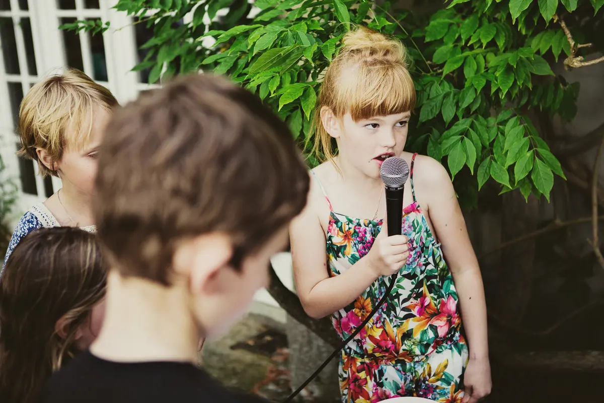 Girl in a floral sundress singing into a microphone as other children watch