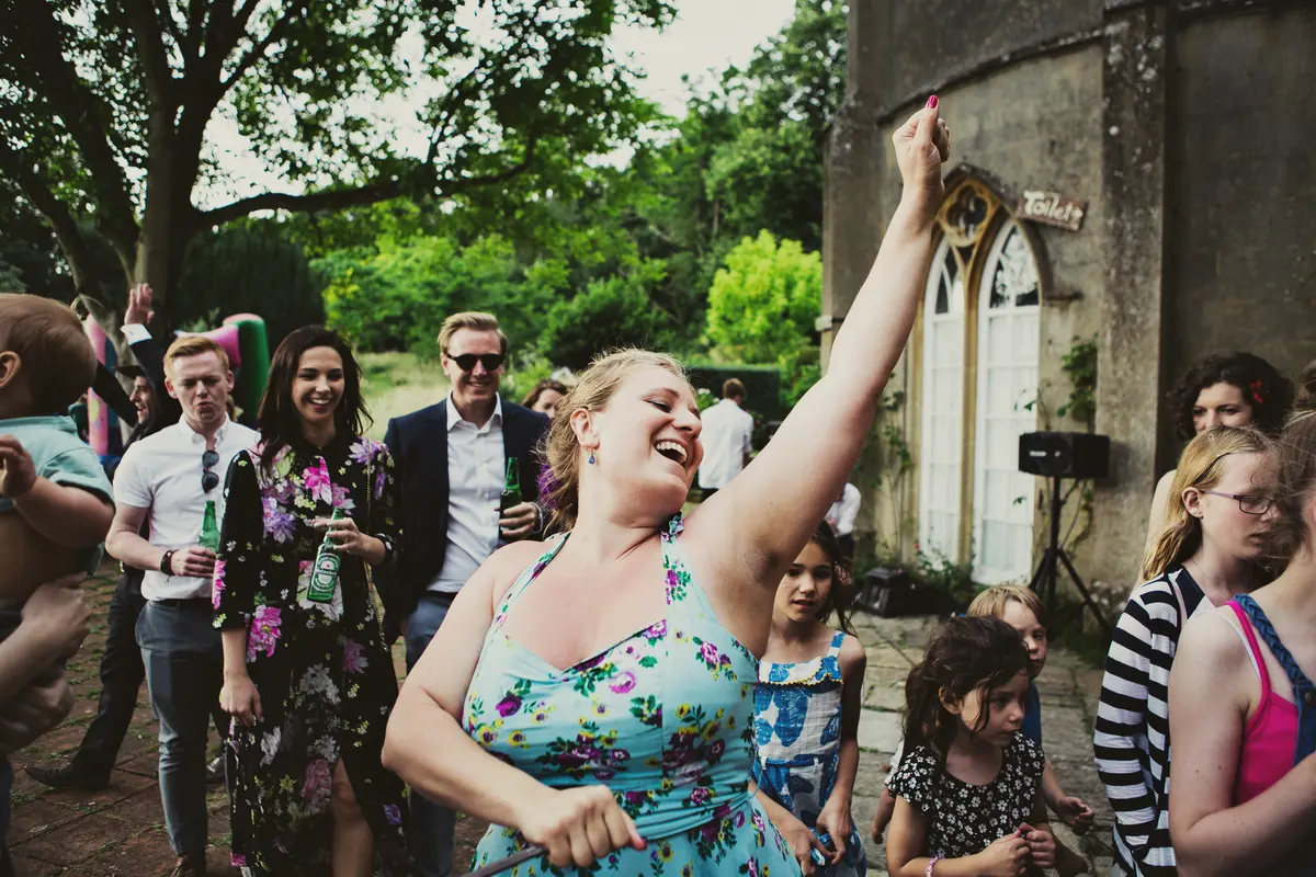 Guest in a floral dress laughing with one arm raised as she dances outside with other guests