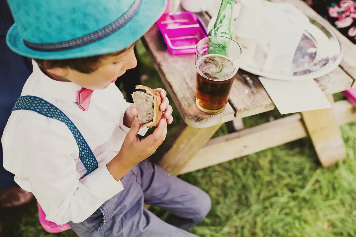 Boy in a trilby, braces and pink bow tie eating a pie at a picnic table