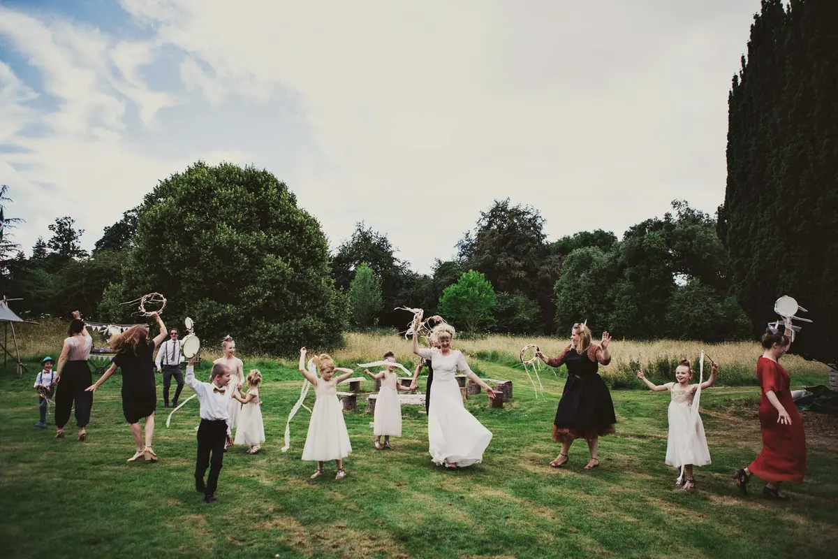 Bride, bridesmaids and flower girls dancing on a lawn with ribboned tambourines