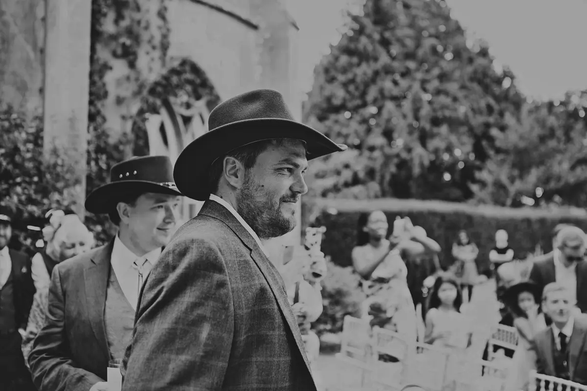 Black and white shot of the groom in a cowboy hat looking back down the aisle as guests stand