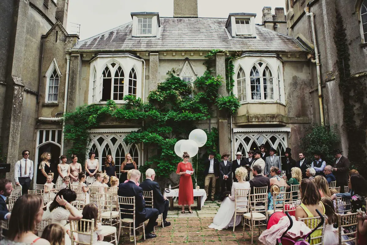 Guests seated in a courtyard of a Gothic house as the celebrant leads an outdoor ceremony beside white balloons