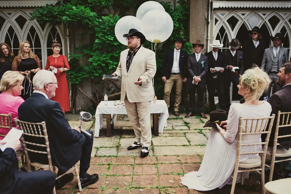 Celebrant in a cream suit and cowboy hat addressing guests at the outdoor ceremony in a courtyard