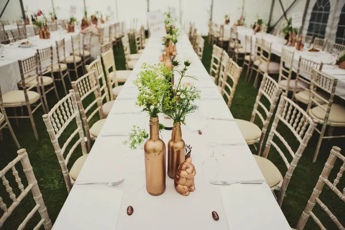 Long tables in a marquee laid with gold bottles of wild flowers and Chiavari chairs