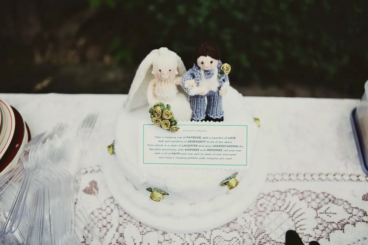 White wedding cake topped with knitted bride and groom figures and a printed recipe for marriage