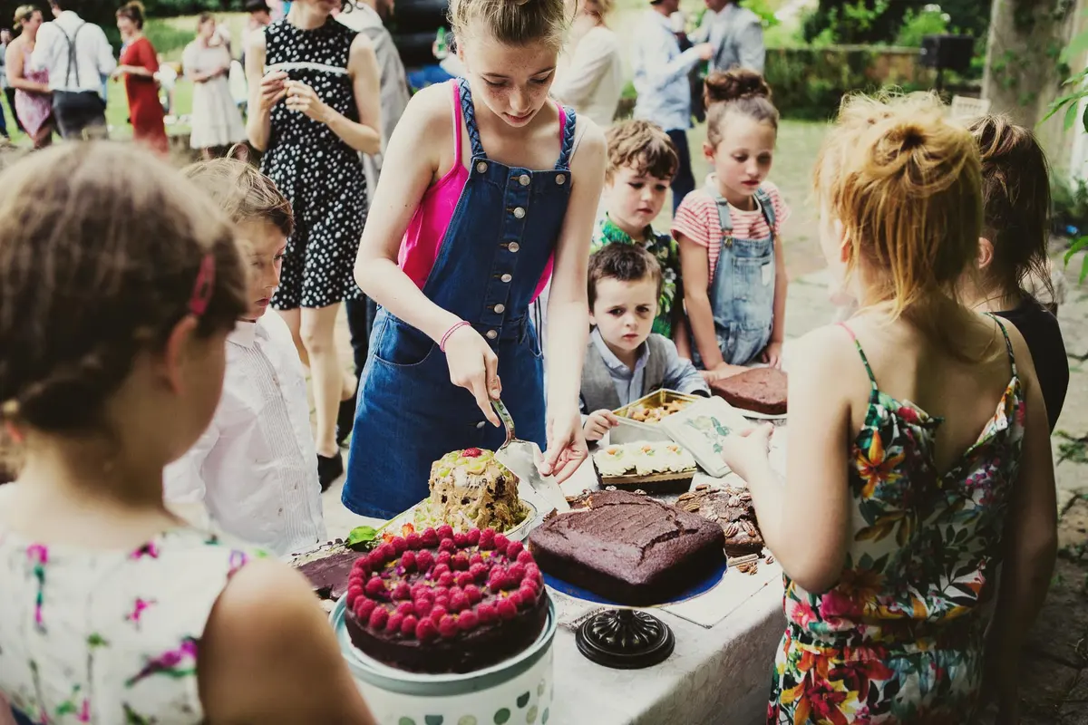 Girl in dungarees cutting cake for a queue of children at an outdoor dessert table
