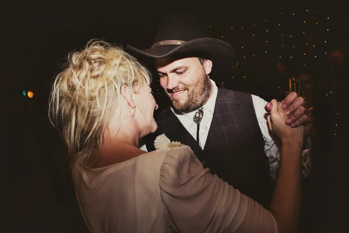 Groom in a cowboy hat and bolo tie smiling as he dances with the bride in the evening