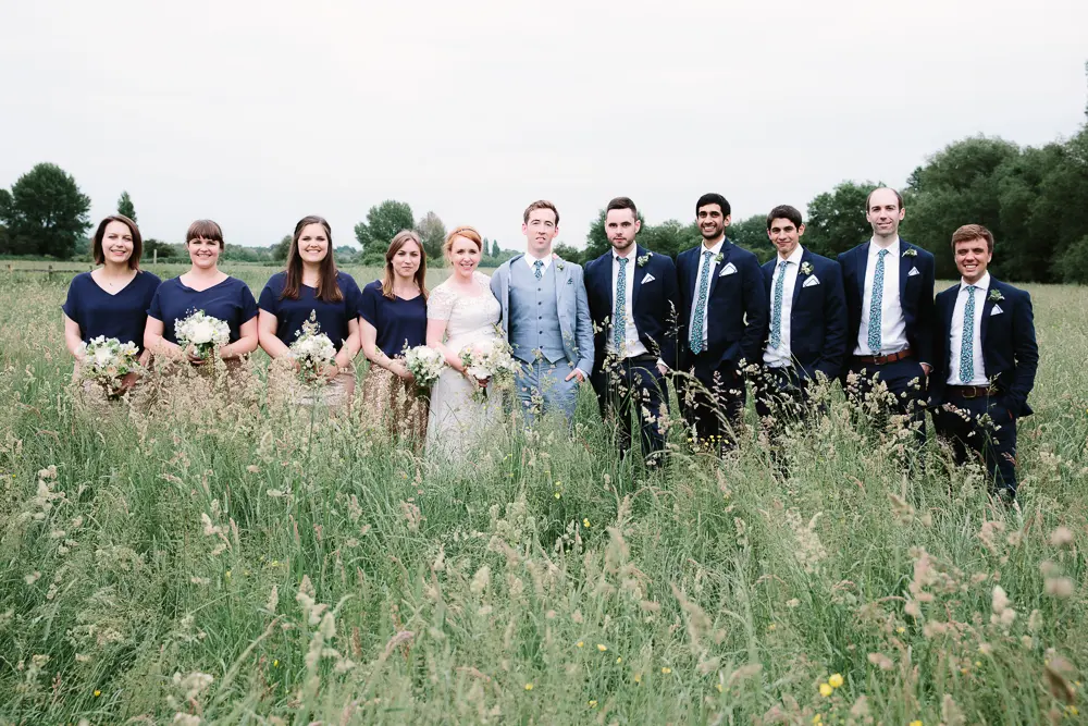 Bride, groom, bridesmaids in navy and groomsmen standing in a meadow of long grass