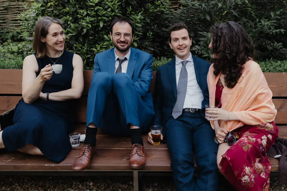 Four guests sitting on a wooden garden bench with drinks, smiling at the camera