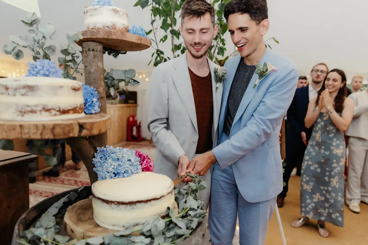 Two grooms cutting a semi-naked cake decorated with blue hydrangeas as guests watch