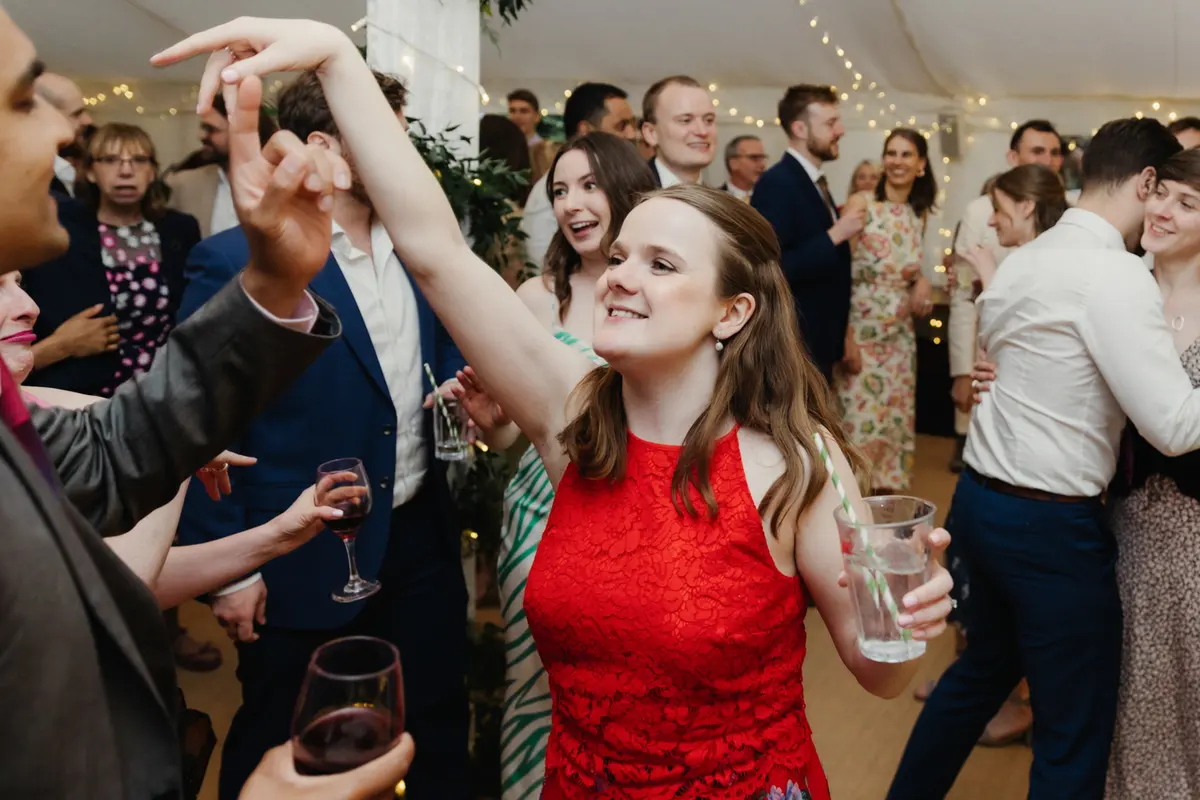 Guest in a red dress dancing with her arm raised among guests in the marquee