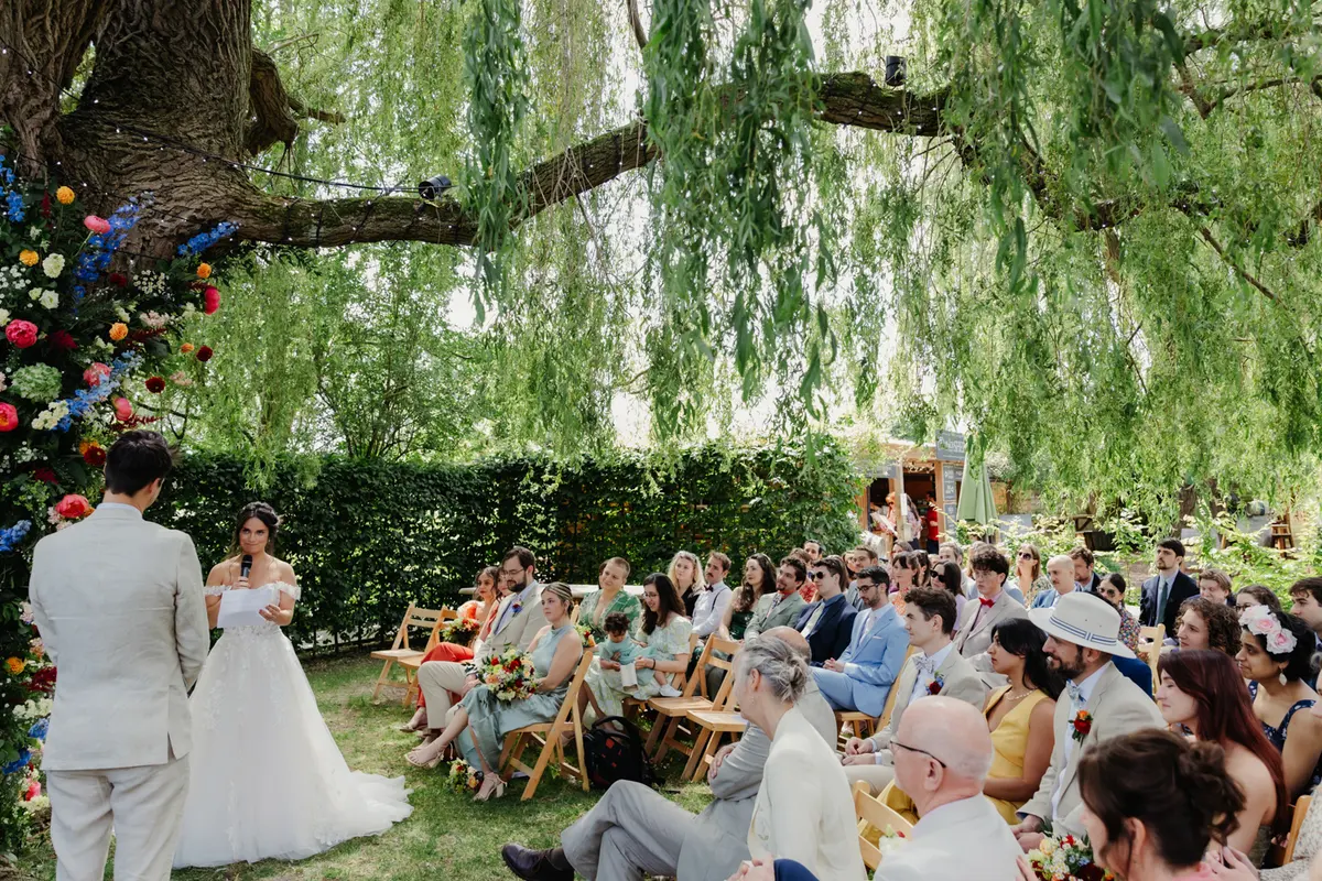 Bride speaking into a microphone at an outdoor ceremony under a willow tree at the Perch