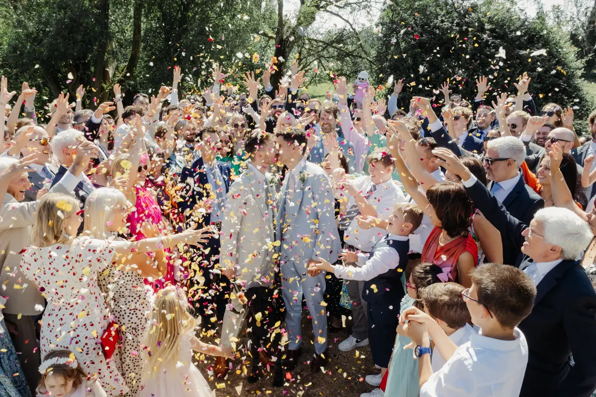 Two grooms kissing as a large crowd of guests throws confetti over them