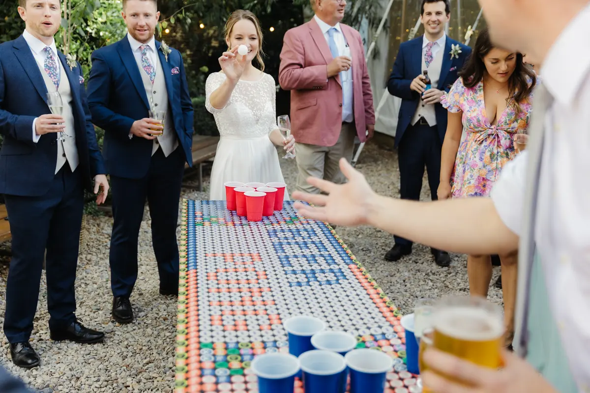 Bride lining up a shot at beer pong as guests in suits watch at the Perch