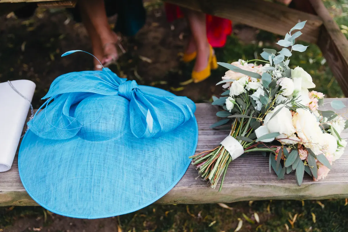 Blue wedding hat and a bouquet of white roses and eucalyptus resting on a wooden bench