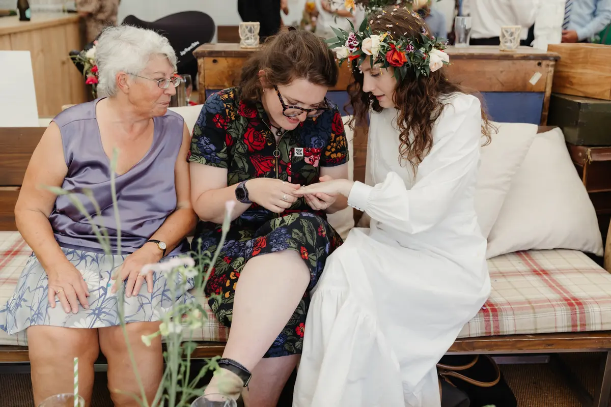 Bride in a flower crown showing her ring to two guests on a sofa in the marquee