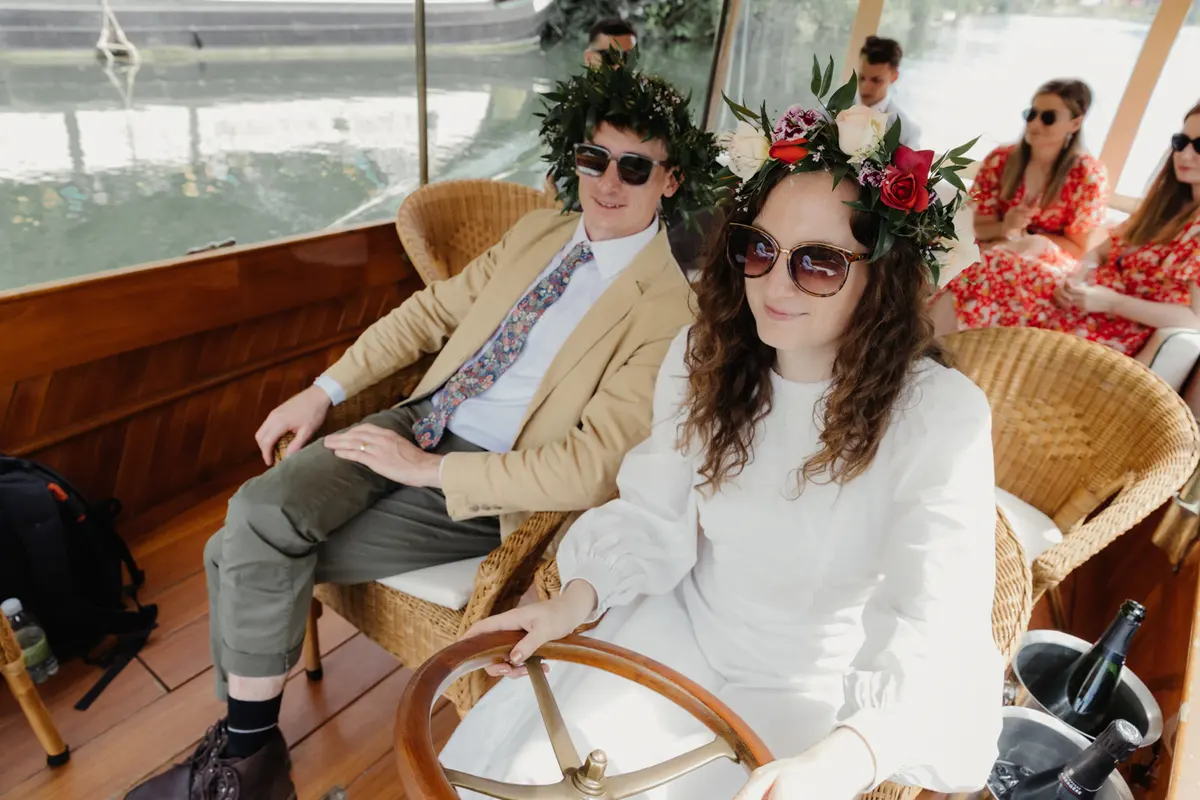 Couple in flower crowns and sunglasses on wicker seats aboard a wooden river boat