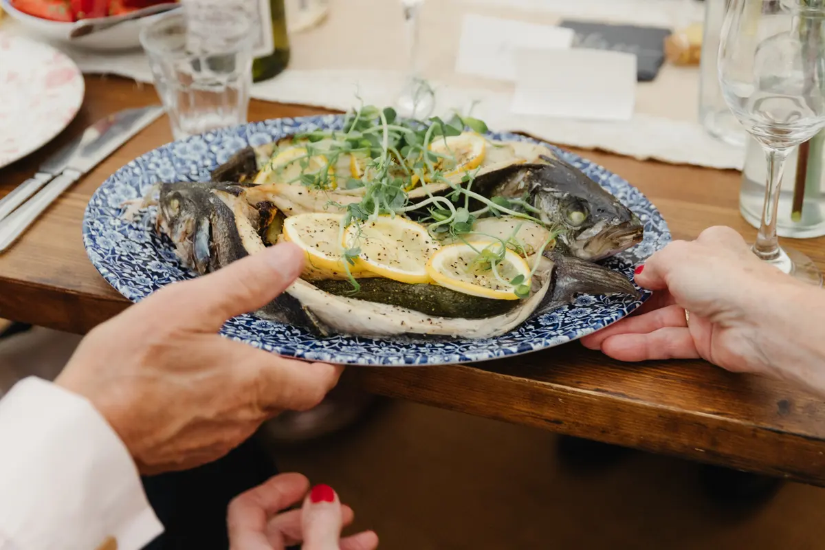 Two guests holding a platter of baked whole fish with lemon slices and herbs