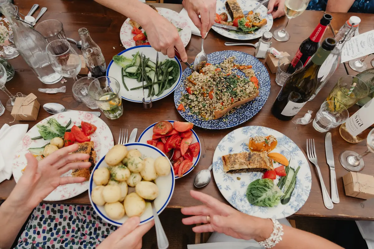 Overhead view of hands reaching for sharing dishes of potatoes, salads and tart