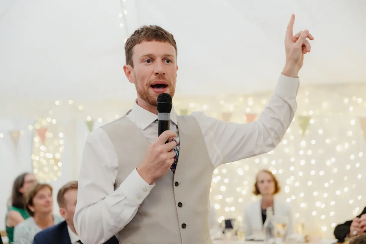 Man in a waistcoat giving a speech with a microphone in front of fairy lights in the marquee