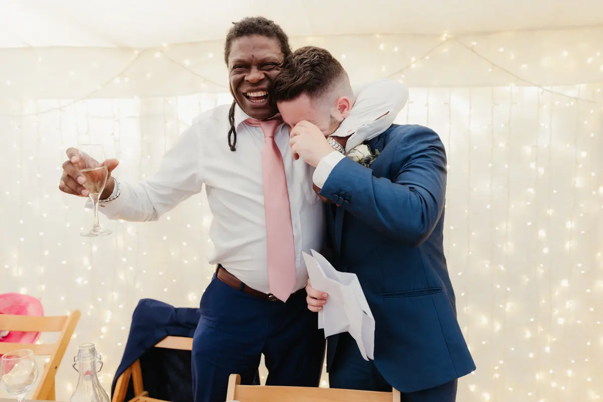 Guest with a glass raised hugging the groom, who wipes away tears after his speech