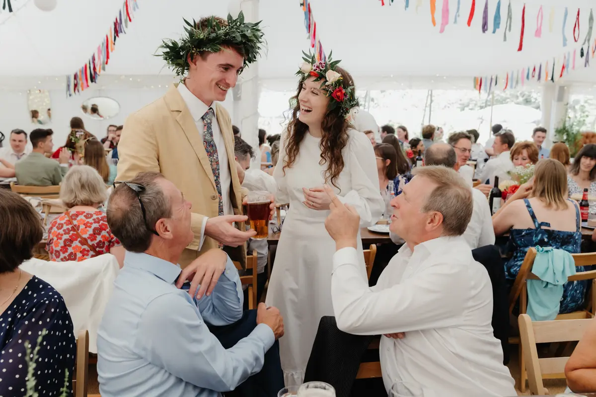 Bride and groom in flower and leaf crowns laughing with guests at the tables in the marquee