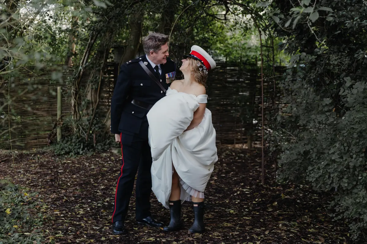 Groom in military dress uniform smiling at the bride, who wears his cap and wellies, in a leafy garden