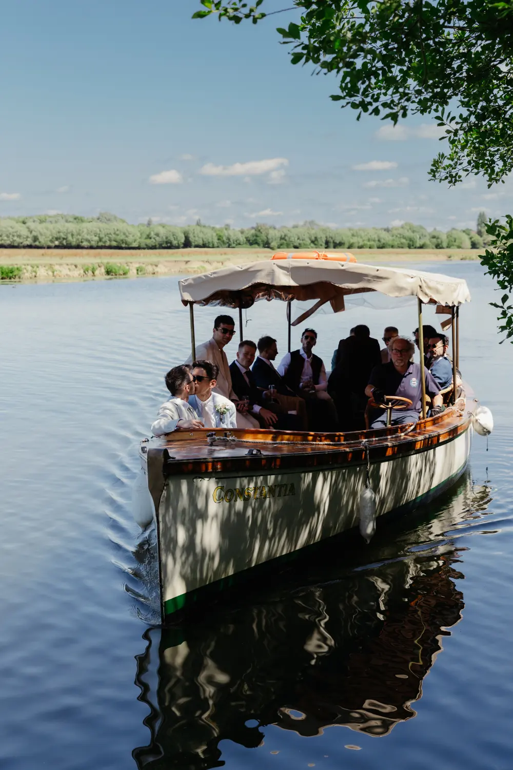 Wedding party arriving by canopied river boat across calm water on a sunny day