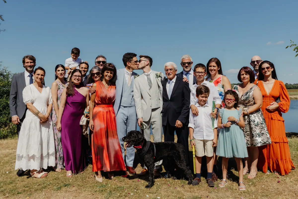Two grooms kissing in a family group photo by the water, with a black dog in front