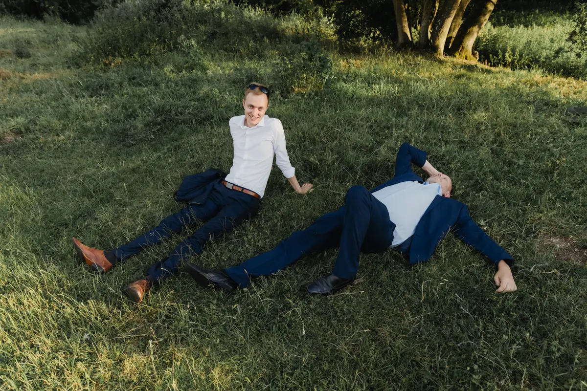 Two men in suits lying on the grass, one sitting up and smiling at the camera