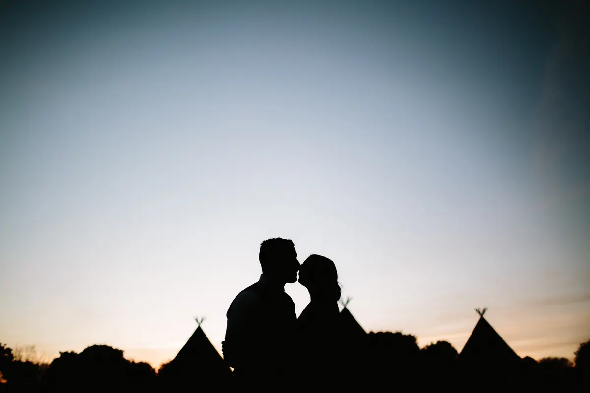 Silhouette of a couple kissing at dusk with tipi tops against the sky