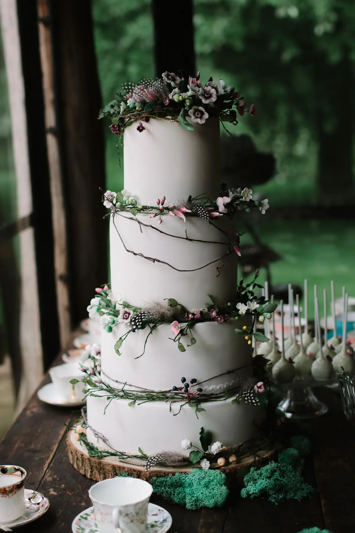 Four tier white wedding cake wrapped in twigs, flowers and feathers beside teacups