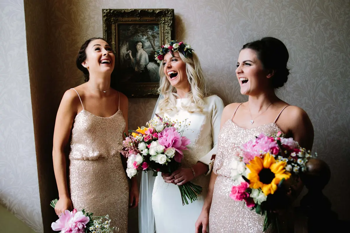 Bride in a flower crown laughing with two bridesmaids in sequinned dresses holding bouquets