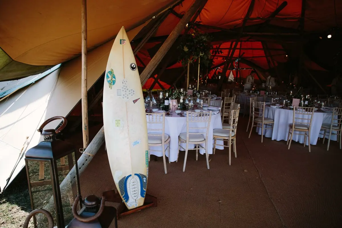 Surfboard propped at the entrance of a tipi set with round tables and white chairs
