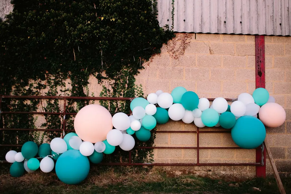 Garland of teal, white and peach balloons along a farm gate by an ivy-covered barn