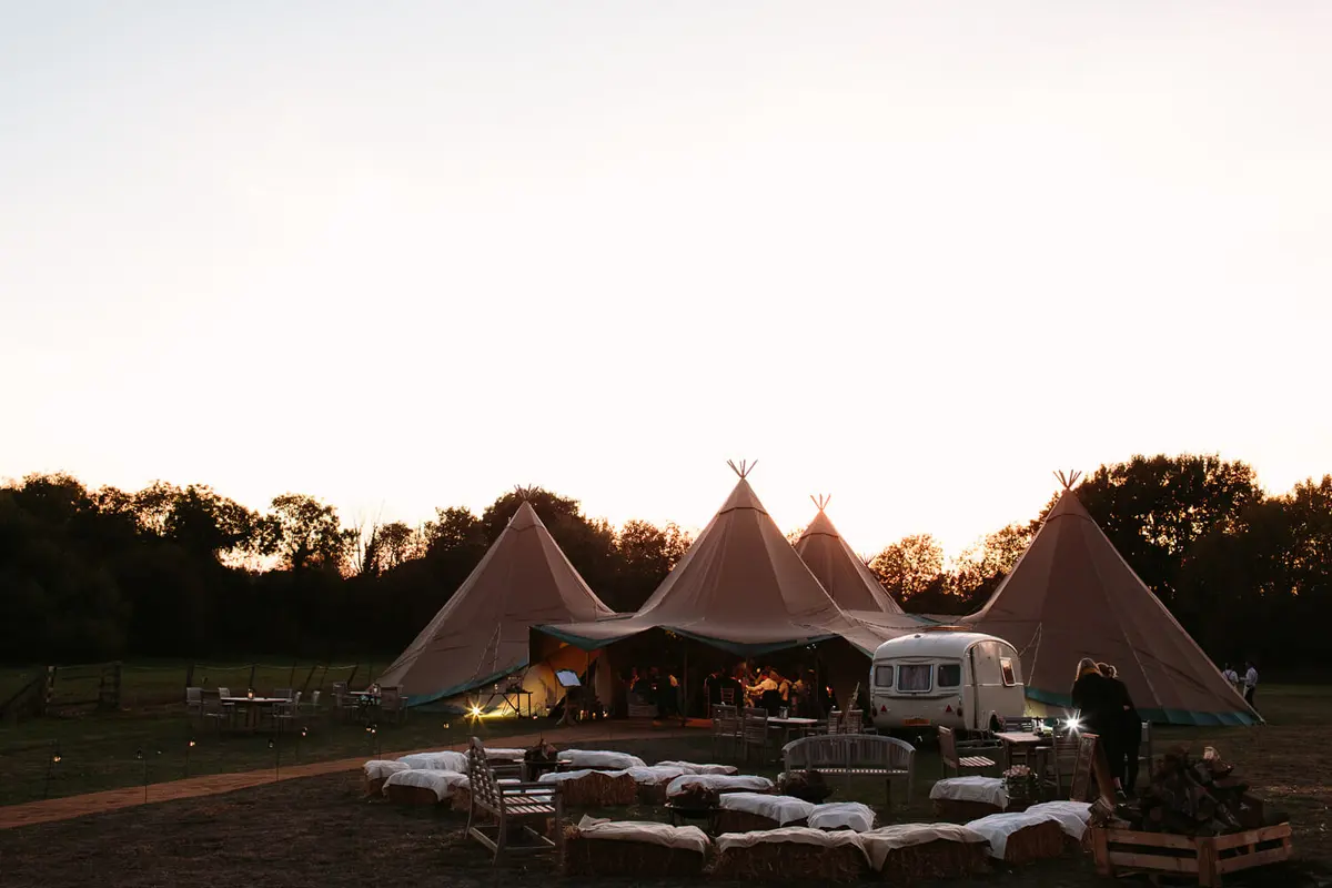 Tipis and a vintage caravan with hay bale seating in a field at dusk