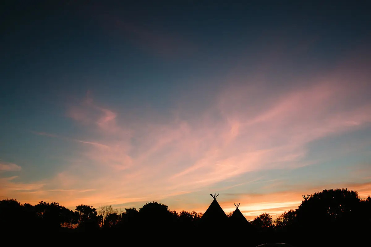 Tipi tops silhouetted against a pink and blue sunset sky over the trees