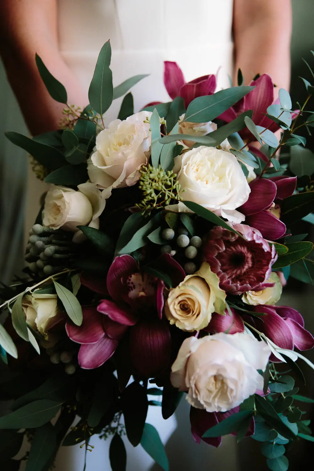 Close-up of a bouquet of white roses, burgundy orchids and eucalyptus held by the bride