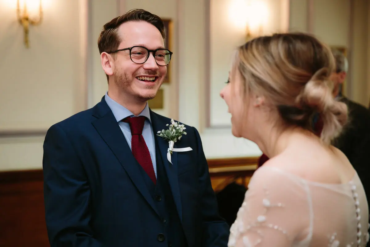 Groom in glasses laughing as he faces the bride during the ceremony