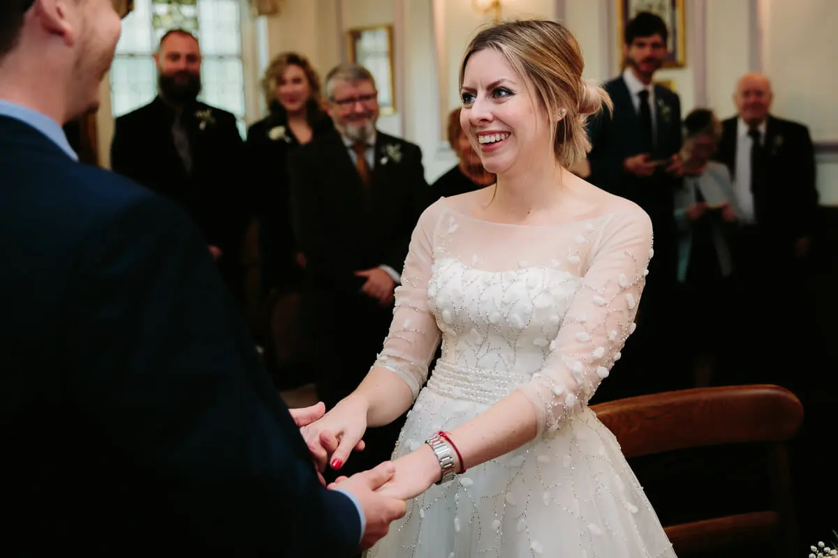 Bride beaming as she holds the groom's hands during the ceremony, with guests behind