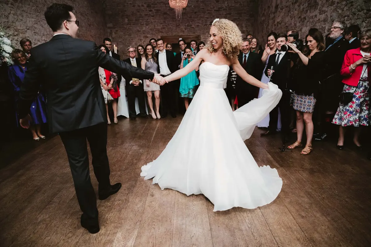 Bride twirling her skirt as she leads the groom onto the dance floor in a stone-walled barn