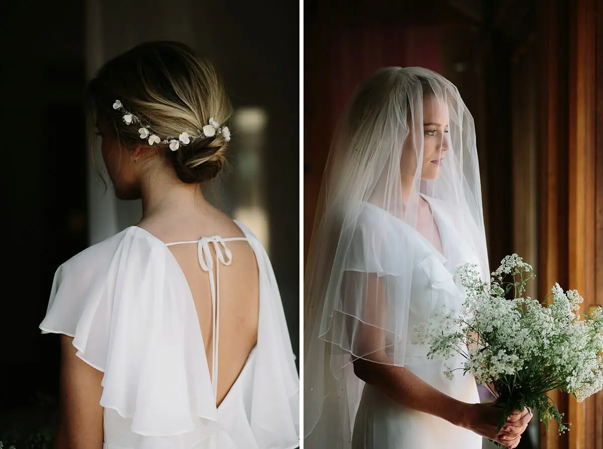 Back of a bride's low bun with small white flowers, and the bride in a veil holding cow parsley by a window